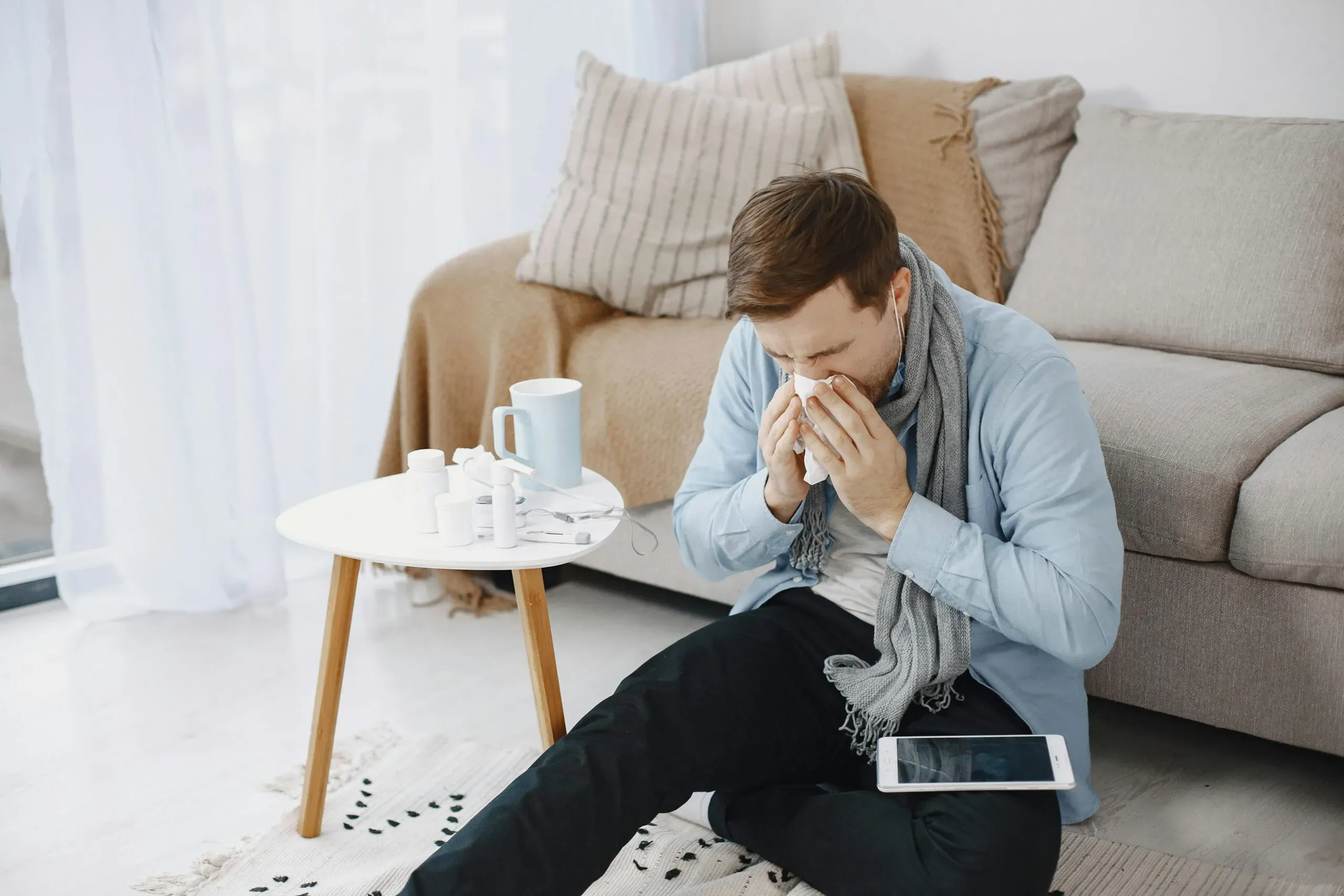 Mann mit Schal sitzt erkältet auf dem Boden vor einem Sofa und putzt sich die Nase